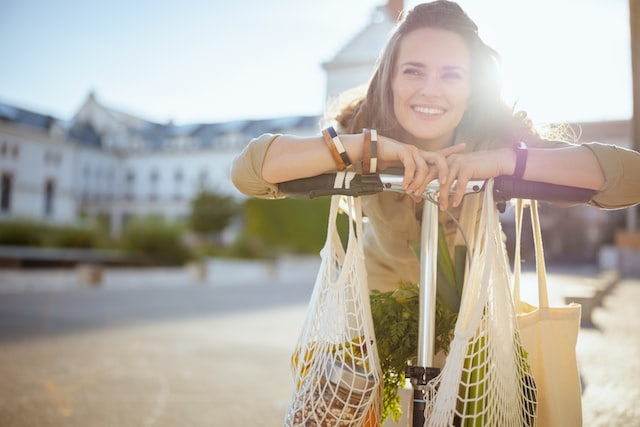 smiling stylish woman in overall with tote bag, string bag and scooter outdoors in the city