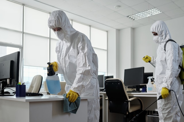 Workers of cleaning service in hazmat suits disinfecting furniture in office